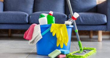 Bucket with sponges, bottles of detergent and mopping stick in front of sofa in living room. Housework cleaning equipment. Cleaning service idea.